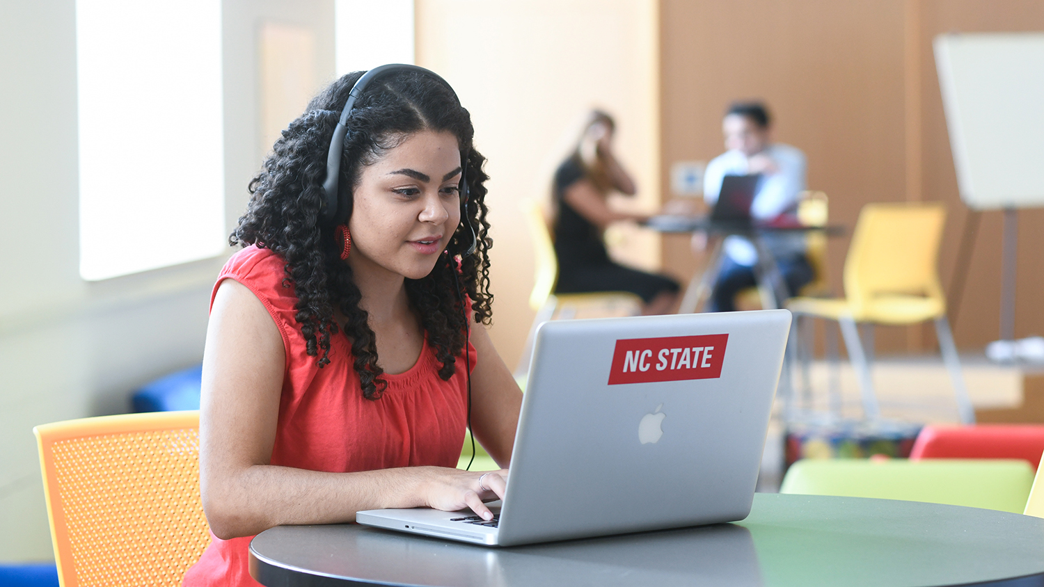 Female student uses computer with headphones in group study space.