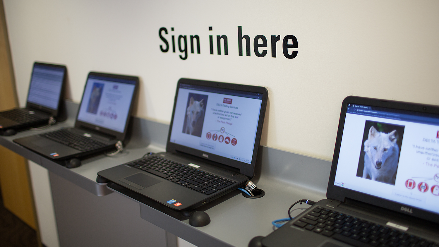 Laptops lined up for students to sign in on. "Sign in here" is written on the wall above.