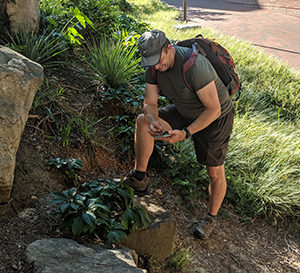 A man leans over looking at his phone next to a large rock.